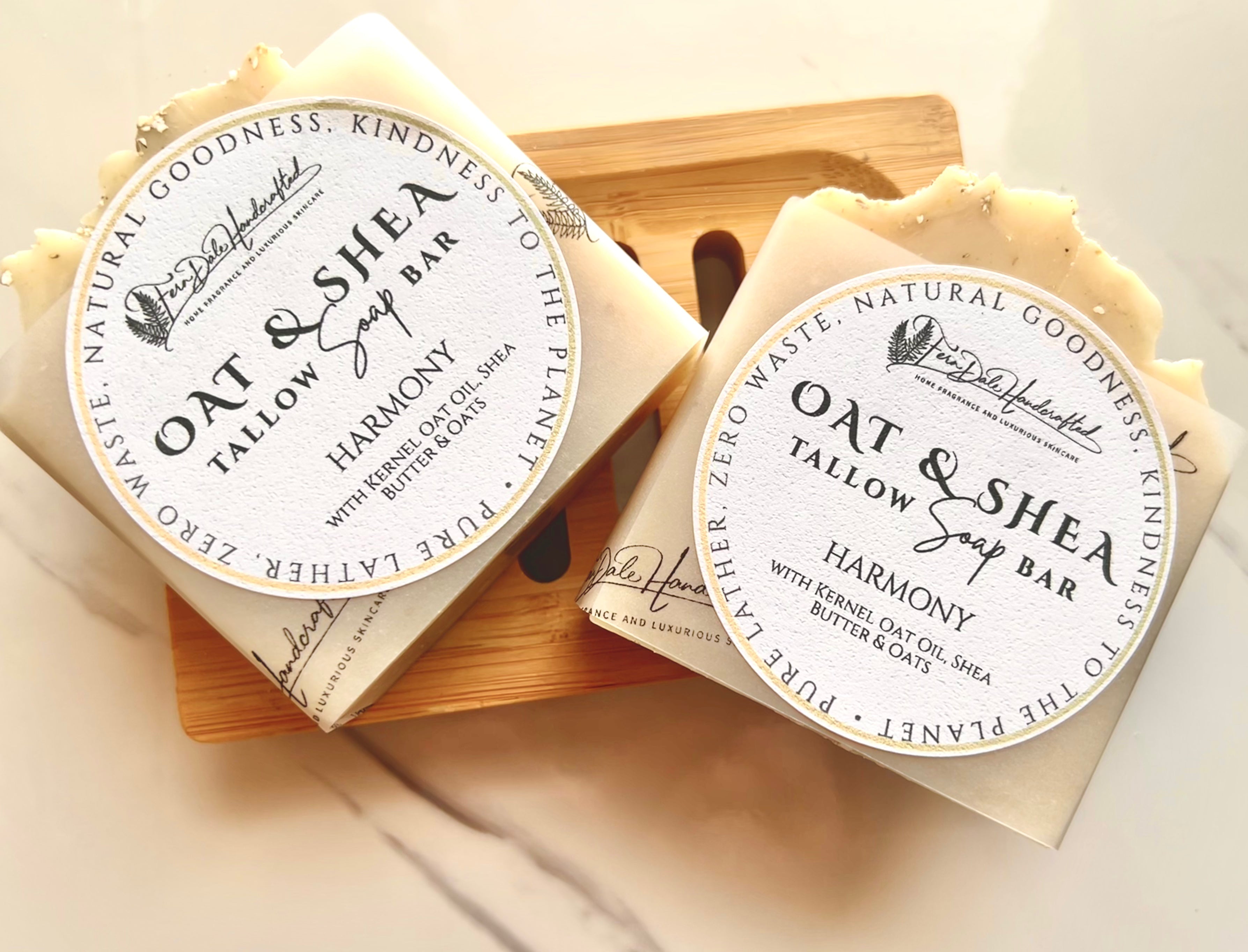 Two cream Oat & Shea Tallow Soap bars with paper bands and round labels rest on a wooden soap dish.