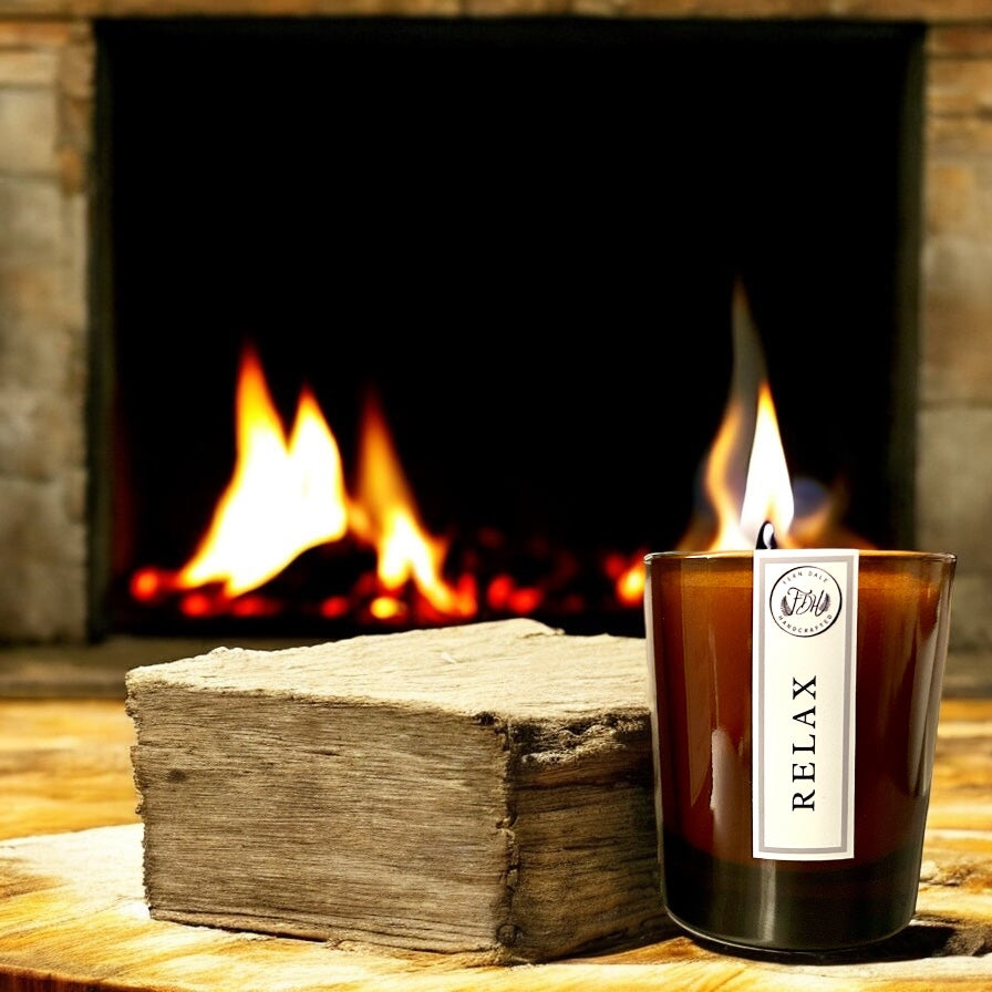 Relax candle burning in an amber glass jar beside a split log, with a lit fireplace behind.