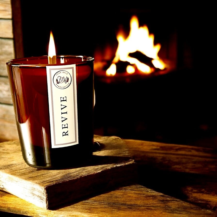 Lit Revive candle in an amber glass jar on a wooden board, with a glowing fire in the background.